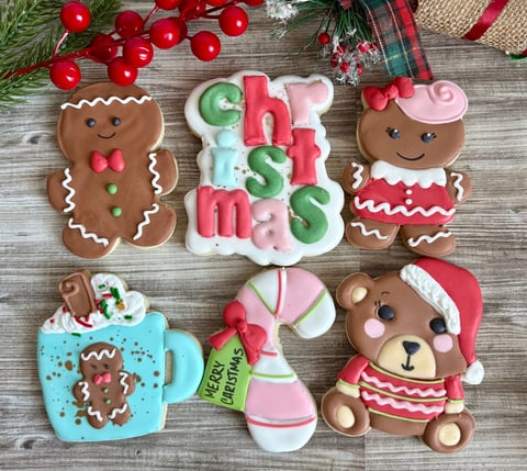 Festive Christmas cookies and decorations arranged on wooden surface, featuring gingerbread people, candy cane, teddy bear, and holiday ornaments in red, green, and pink