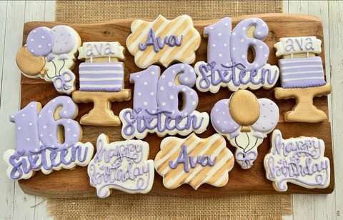 Decorative cookies on a wooden board spelling Ava is 6 with lavender, gold, and white frosting designs
