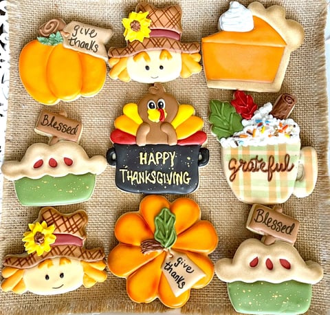 Decorative Thanksgiving-themed sugar cookies arranged on burlap, featuring pumpkins, turkeys, pies, and autumn leaves with text like Happy Thanksgiving and Grateful