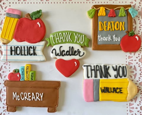 Decorated sugar cookies spelling thank you with teacher-themed designs including apples, pencils, books, and a desk on a polka dot background