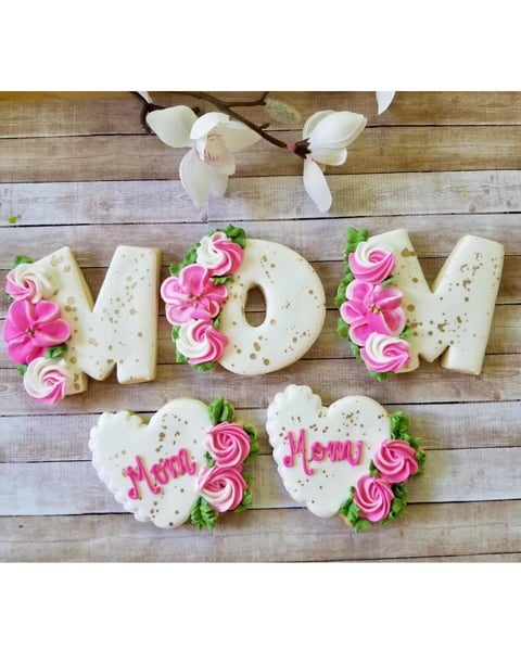 Decorated cookie display spelling MOM and MENU with pink roses, heart shapes, and white orchids on wooden background