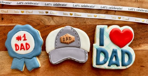 Three Fathers Day decorated cookies: blue ribbon medal with #1 Dad, gray DAD cap, and I love Dad with red heart on wooden surface with celebratory ribbon
