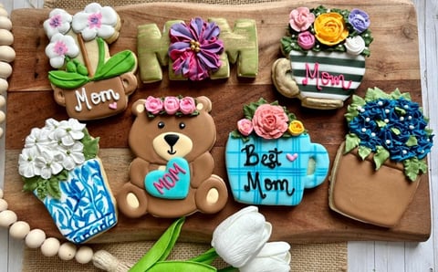 Decorated cookies featuring Mom themed designs with flowers, teddy bear, and potted plants on a wooden board