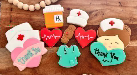 Decorated cookies themed for healthcare workers, including pills, stethoscopes, hearts with heartbeat lines, nurse caps, and thank you messages on a wooden surface