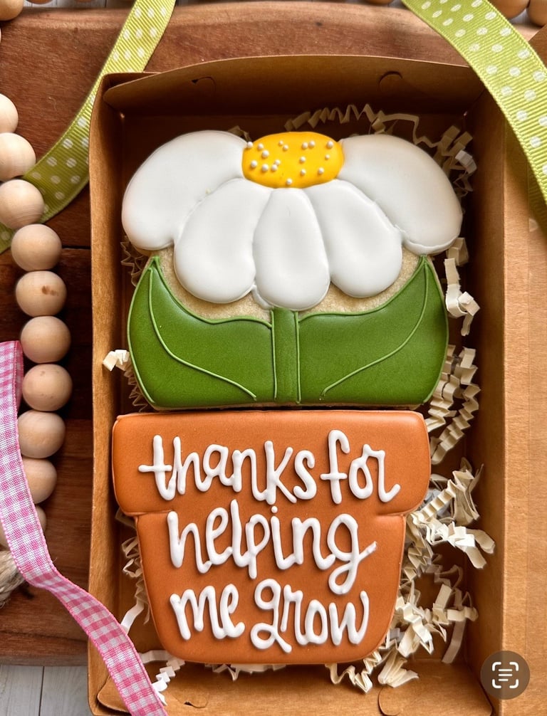Decorated sugar cookies shaped as a white daisy flower and orange pot with thanks for helping me grow text, arranged in a cardboard box with ribbon