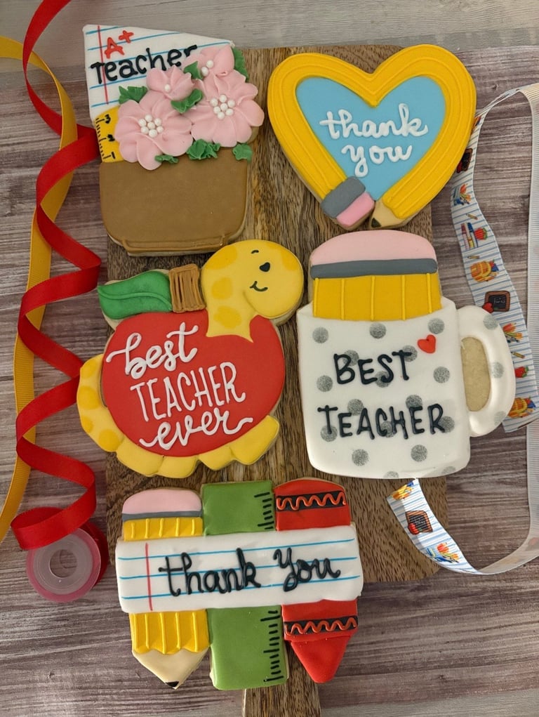 Decorative teacher appreciation cookies including designs for thank you, best teacher, and flowers arranged on a wooden surface with red ribbon